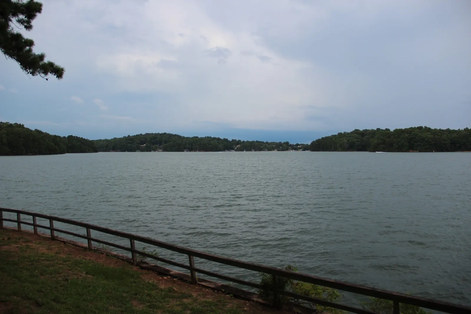 Wide view over the Baldridge Creek arm of Lake Lanier under a heavy grey summer sky: choppy grey-green water, wooded shorelines closing in from both sides with a few lake houses and docks tiny on the far shore, a mown grass bank and a rustic wooden split-rail fence along the bottom right, a pine bough intruding at top left. No people, no signs.