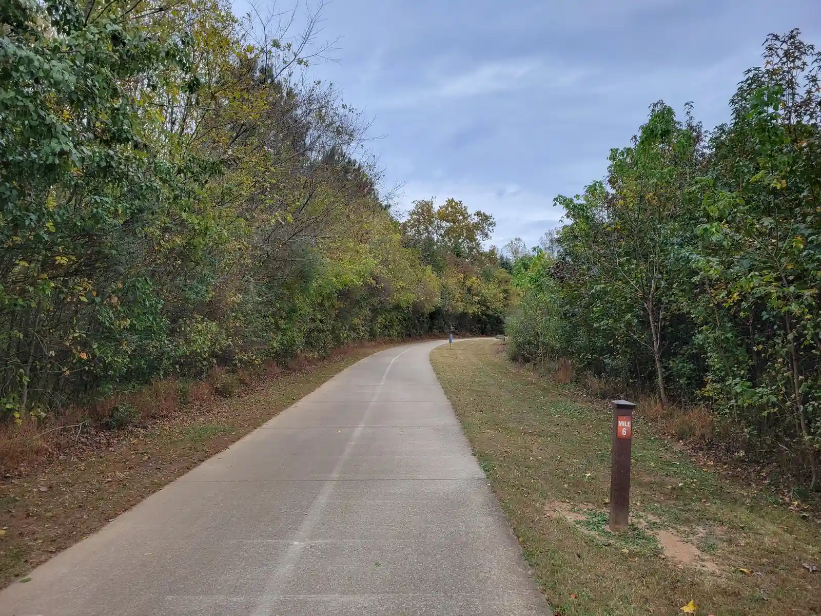 Concrete multi-use path curving away under an overcast autumn sky between thickets of green and yellowing trees; dry grass verges, a brown 'MILE 6' post at right; one distant cyclist/walker far down the path, too small to identify.
