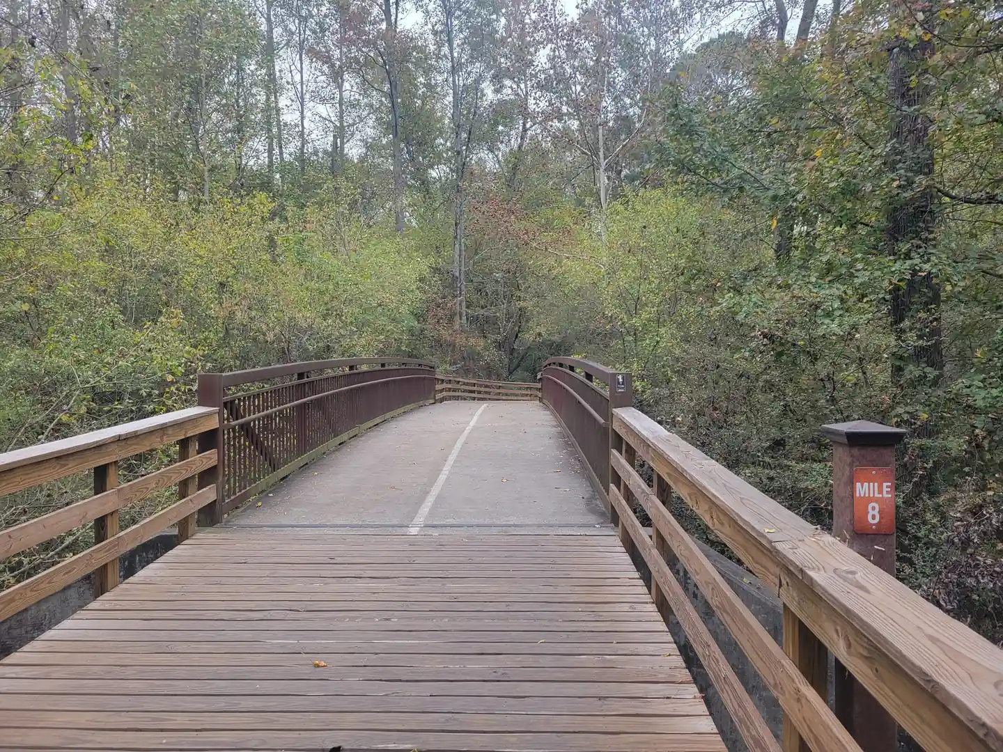 Low-angle view along a timber-decked pedestrian bridge with brown steel truss rails and a painted centre line, a brown post with an orange 'MILE 8' plate at right, thick autumn-tinged woods ahead under a grey sky. No people.