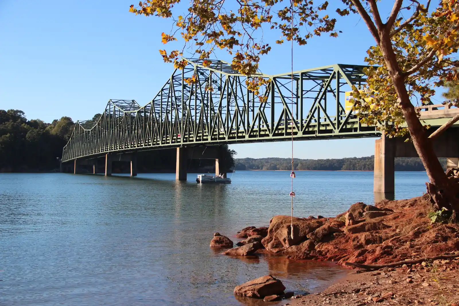 Late-afternoon autumn light on the long green steel truss of Browns Bridge stretching from the left across blue Lake Lanier; a small pontoon boat under the span, a rope swing hanging from a leaning sycamore with golden leaves at right, red Georgia-clay rocks and roots at the water's edge in the foreground, wooded far shore. No people visible, no signs legible.