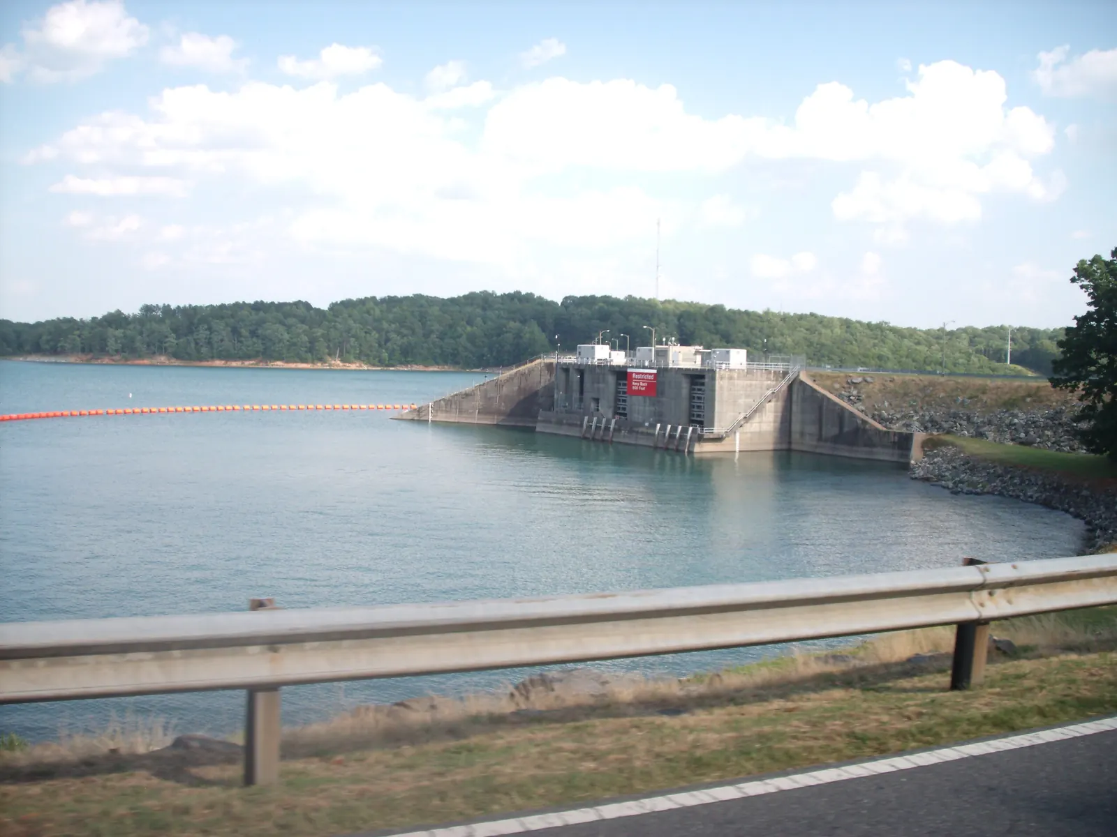 Lake Lanier at the Buford Dam powerhouse intake, shot through a car window: a metal guardrail and road shoulder across the bottom, a row of orange safety buoys across the water, the concrete intake structure with a red 'Restricted' notice, rip-rap embankment at right, wooded far shore and cumulus sky. Slight motion blur on the guardrail. No people.