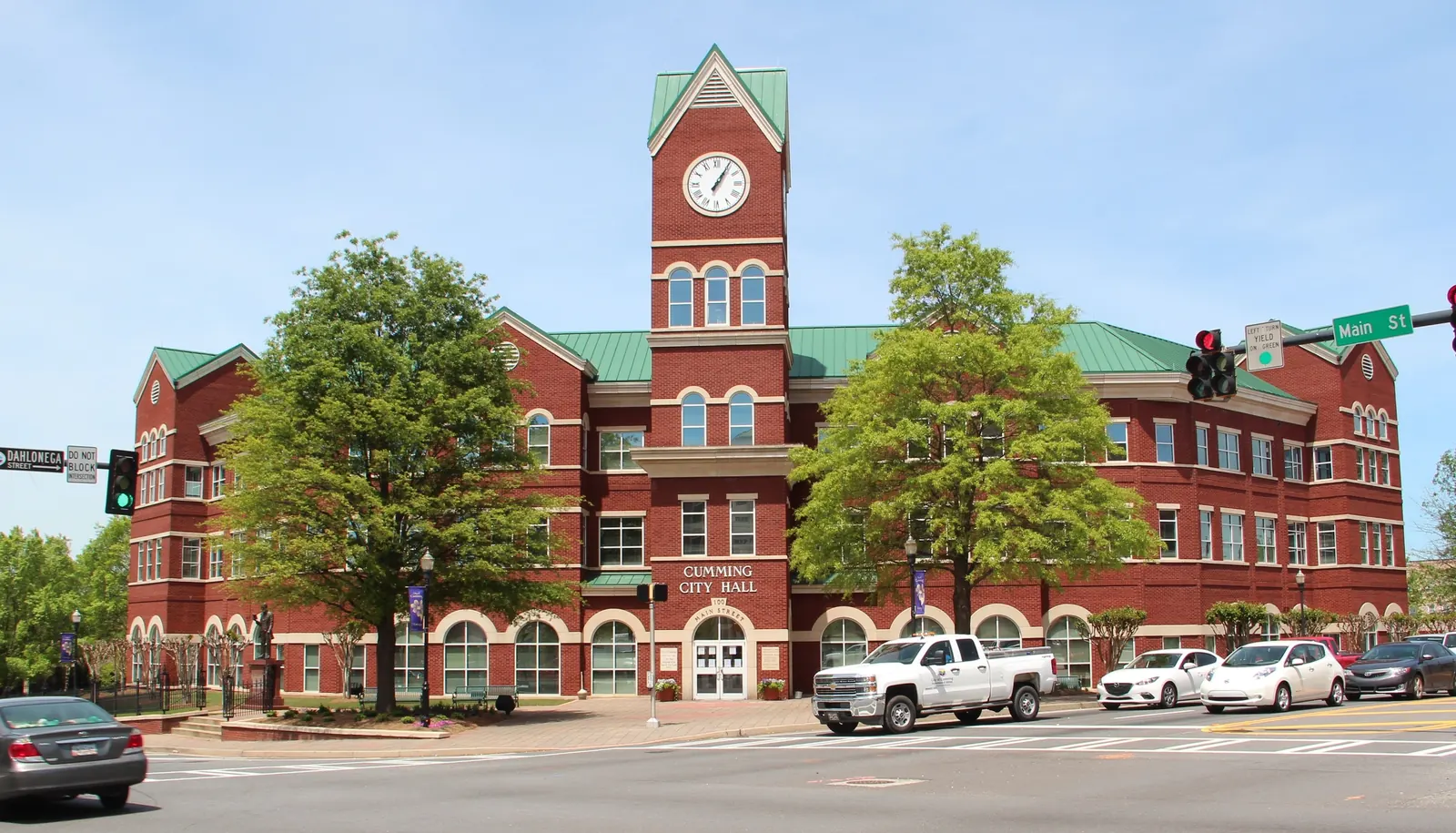 Spring street view of the three-storey red-brick Cumming City Hall with a central clock tower, green standing-seam roofs and arched ground-floor windows, 'CUMMING CITY HALL' lettering over the entrance, two young trees out front; street signs 'Main St' and 'Dahlonega Street' and a traffic signal at right; a white pickup and several sedans parked/passing in the foreground. No people prominent.