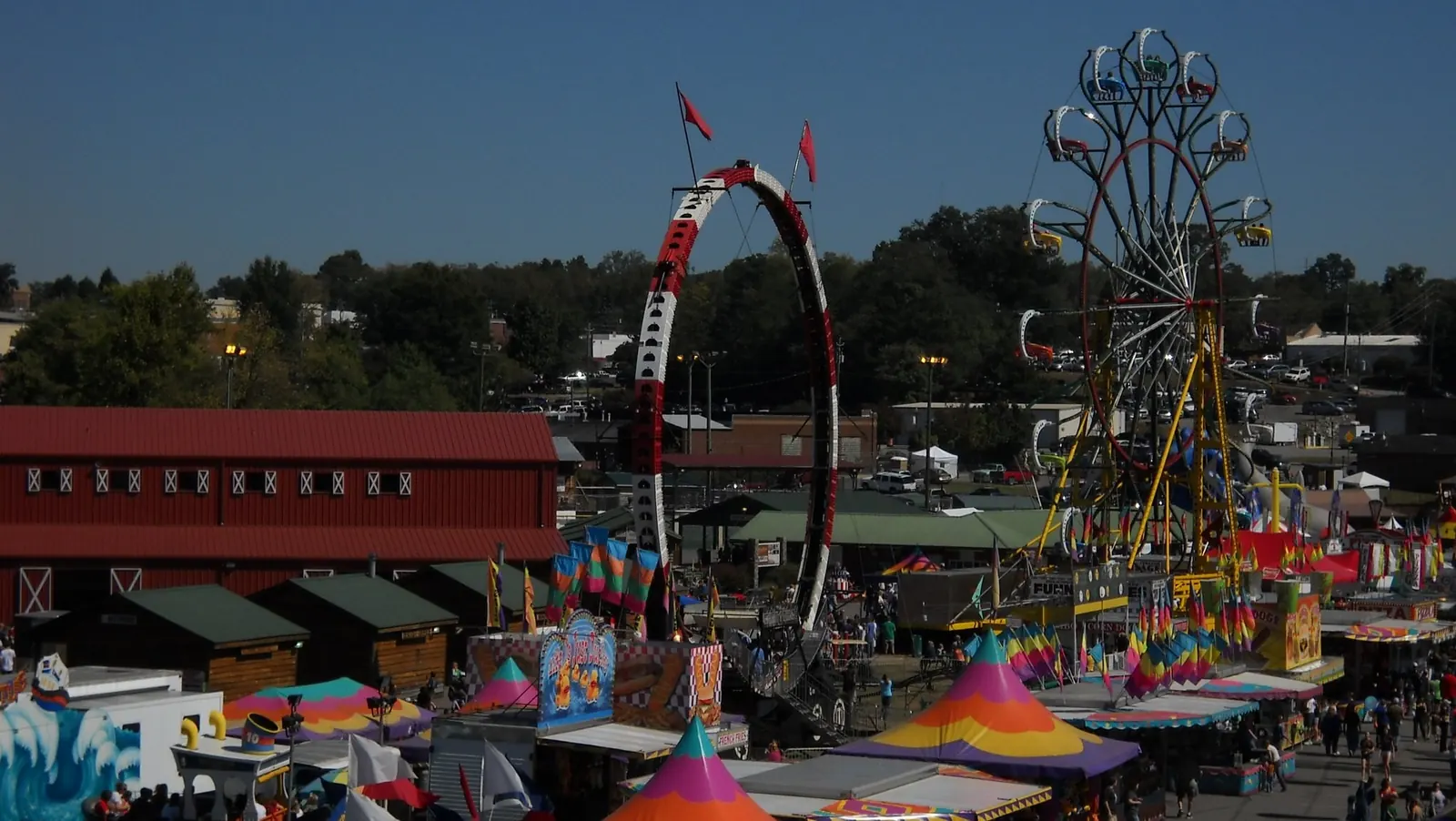 Elevated view over the fair midway on a sunny October day: a long red barn-style exhibit hall with white X-braced windows at left, a tall red-and-white loop ride and a Ferris wheel, colourful striped tents, flags and food stands ('FUNNEL ...', '... DOGS' concession signs), small crowds walking the lanes, parked cars and town buildings beyond under a deep blue sky.