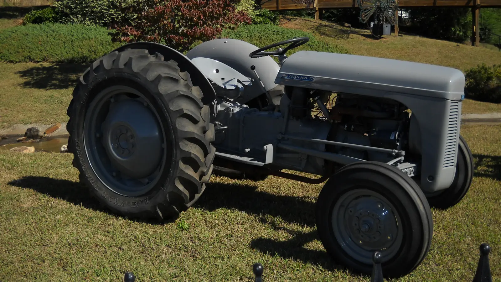 A restored grey Ferguson 30 tractor parked on a sunny lawn behind a black iron fence (finials along the bottom edge), shrubs and a wooden boardwalk/fair building behind. The only lettering is the 'Ferguson' maker's badge on the hood. No people.