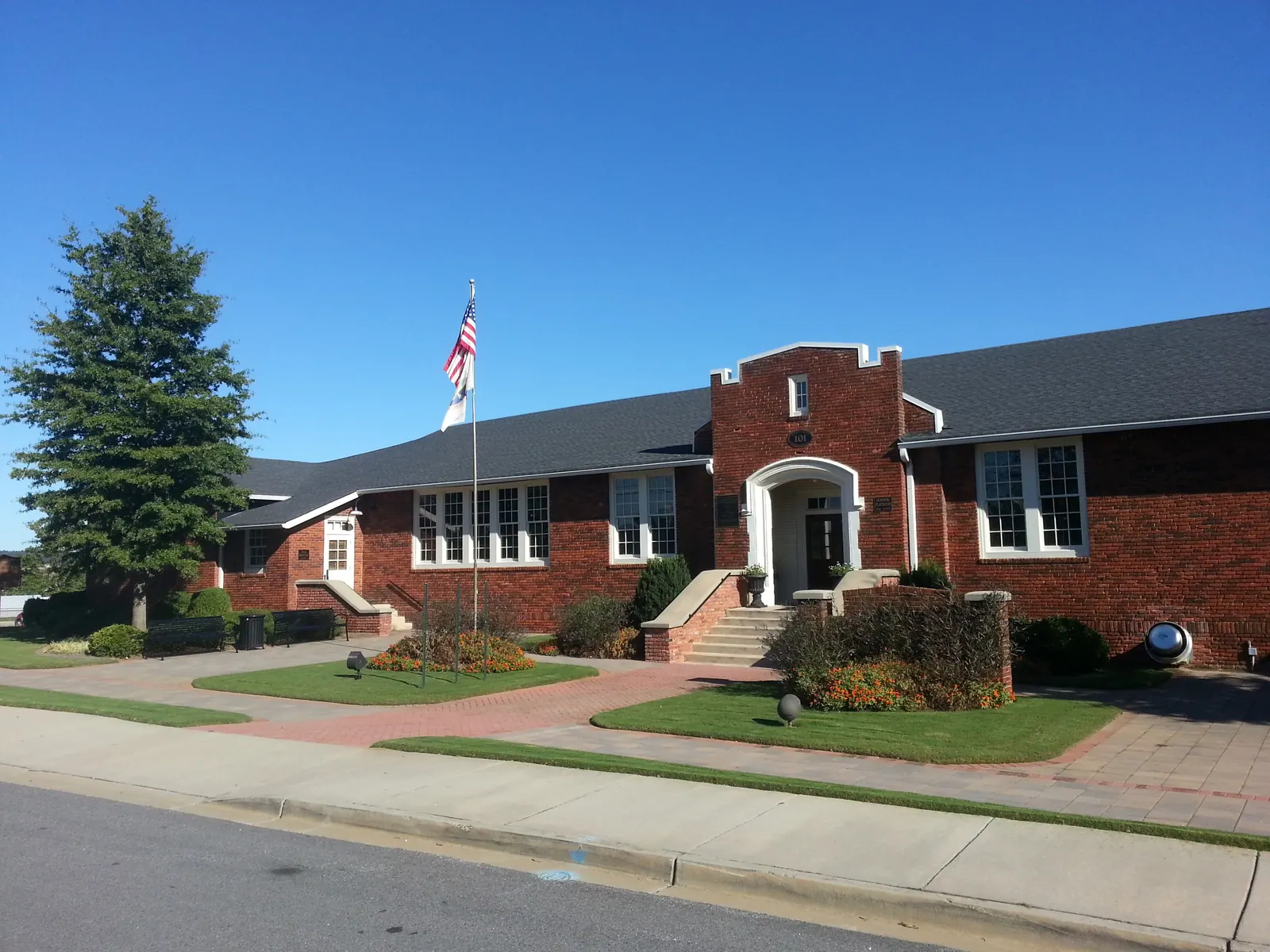 Single-storey 1920s red-brick school building with a crenellated central entrance bay, arched doorway and tall multi-pane windows, a flagpole with the US flag, a tall cedar at left, neat lawns, brick walkways and flower beds, an empty street in front under a deep blue sky. No people; only the house number '101' is legible.