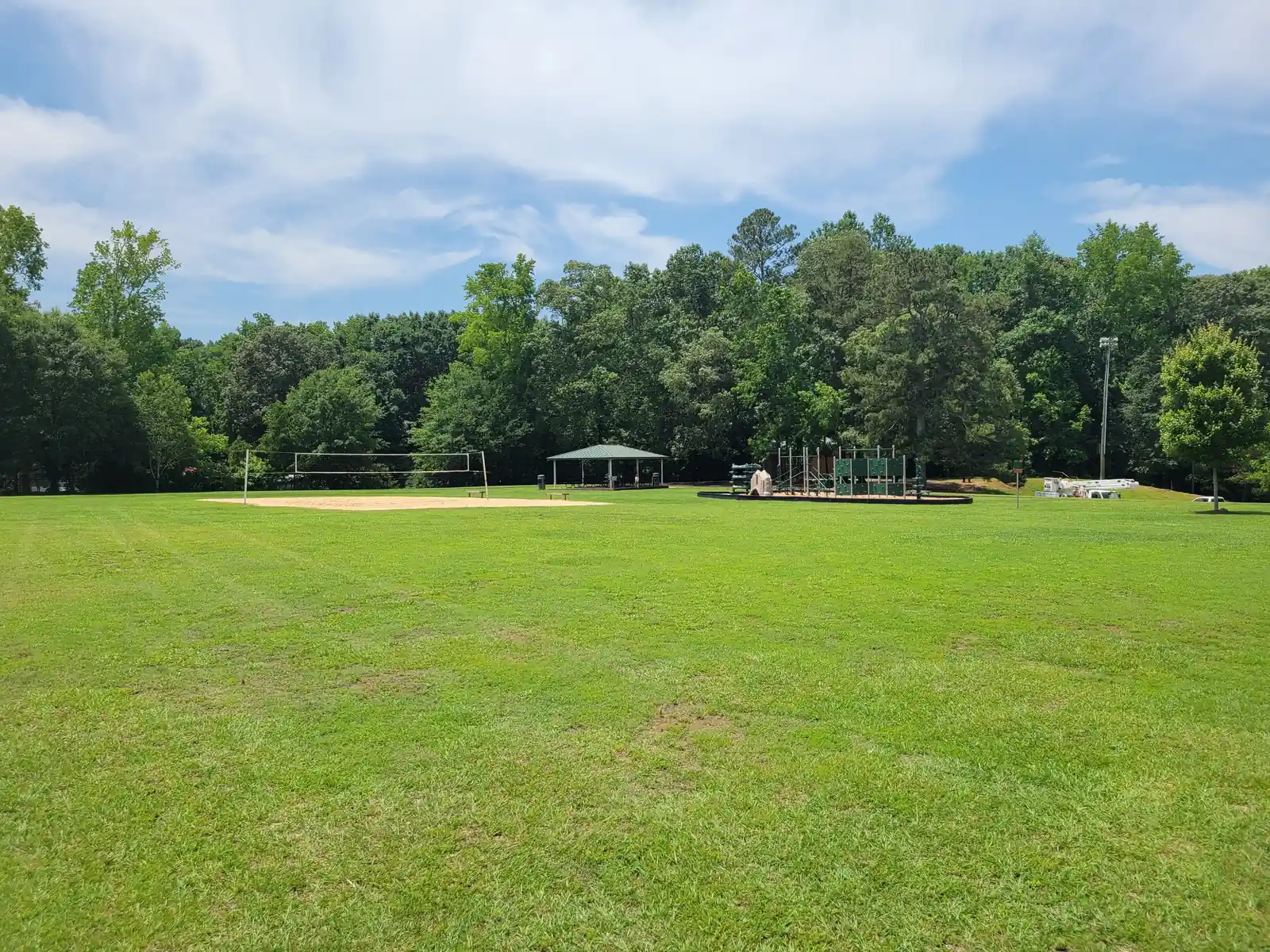 Wide summer view across a large mown lawn to a sand volleyball court, a green-roofed picnic pavilion and a playground structure, with a tall tree line and a white-clouded blue sky above; one adult sitting far away near the playground, too small to identify.