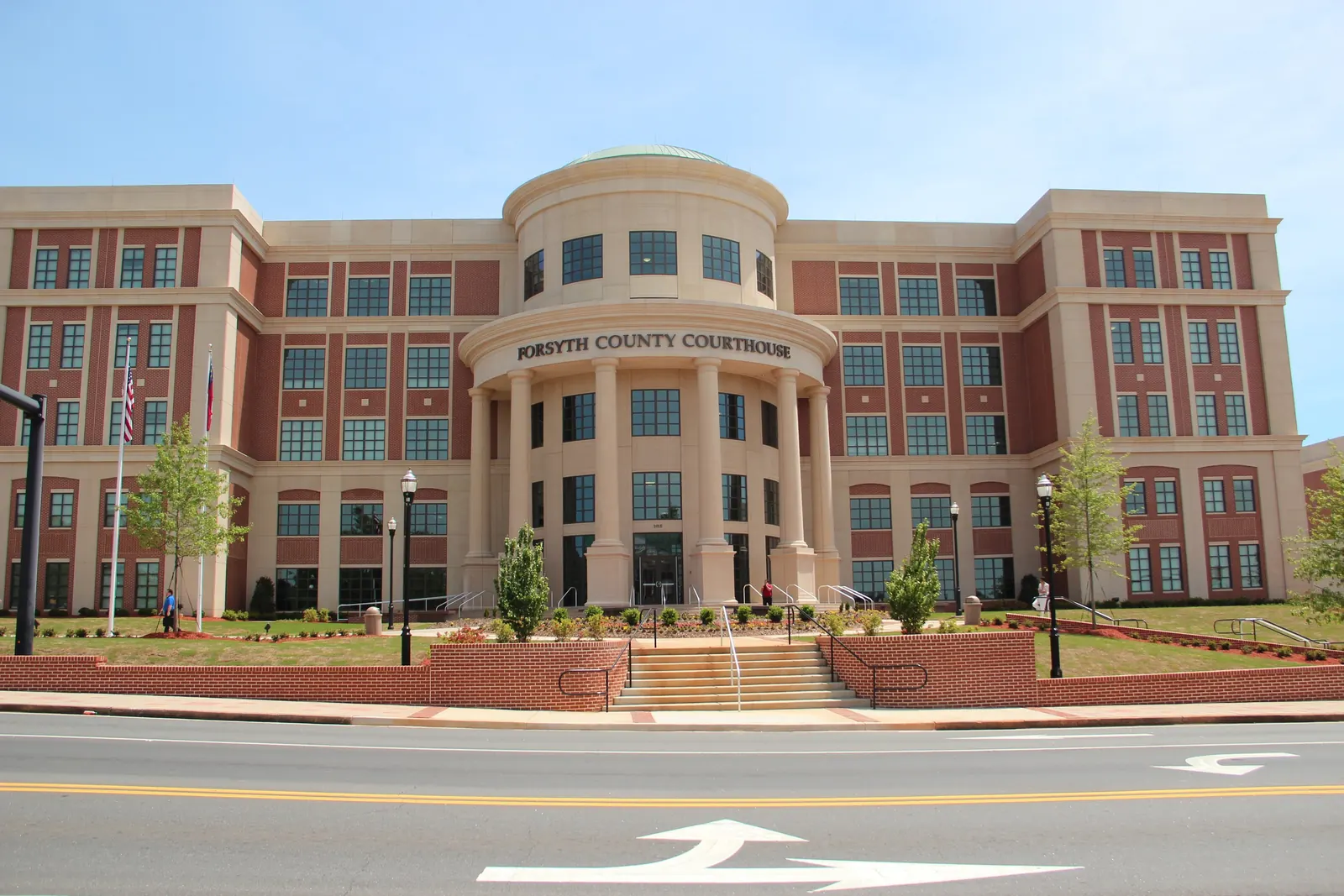 Frontal view of the five-storey brick-and-limestone Forsyth County Courthouse with a domed rotunda and columned portico lettered 'FORSYTH COUNTY COURTHOUSE', US and Georgia flags at left, landscaped brick terraces and steps, and an empty street with turn arrows in the foreground. A couple of distant pedestrians, not identifiable.
