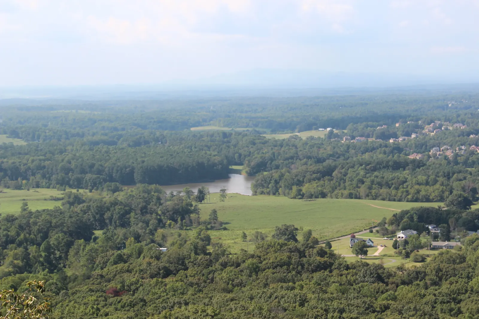 Telephoto view down from Sawnee Mountain over rural Forsyth County: dense green woods in the foreground, a curving pond, open hay pastures with a dirt track, a cluster of farmhouses with outbuildings, and newer subdivision rooftops in the middle distance, fading into hazy ridges under a washed-out white sky.