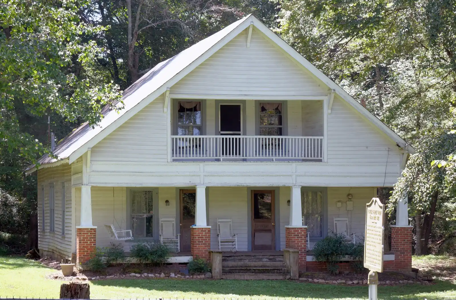 Front view of a white two-storey clapboard farmhouse with a steep gable, an upper recessed balcony with white balustrade, a full-width front porch on brick piers with rocking chairs and a porch swing, shade trees around it, and a historical marker sign on a post at right (text not legible at this size). No people.