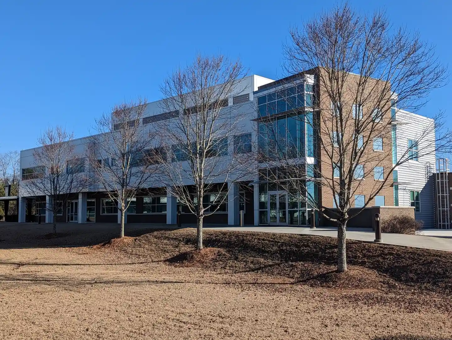 Three-storey modern campus building with white metal panels, a tan brick stair tower and a full-height glass corner, a row of bare young trees on a mulched berm in front, dormant brown lawn and a clear blue sky. No people, no legible signs.