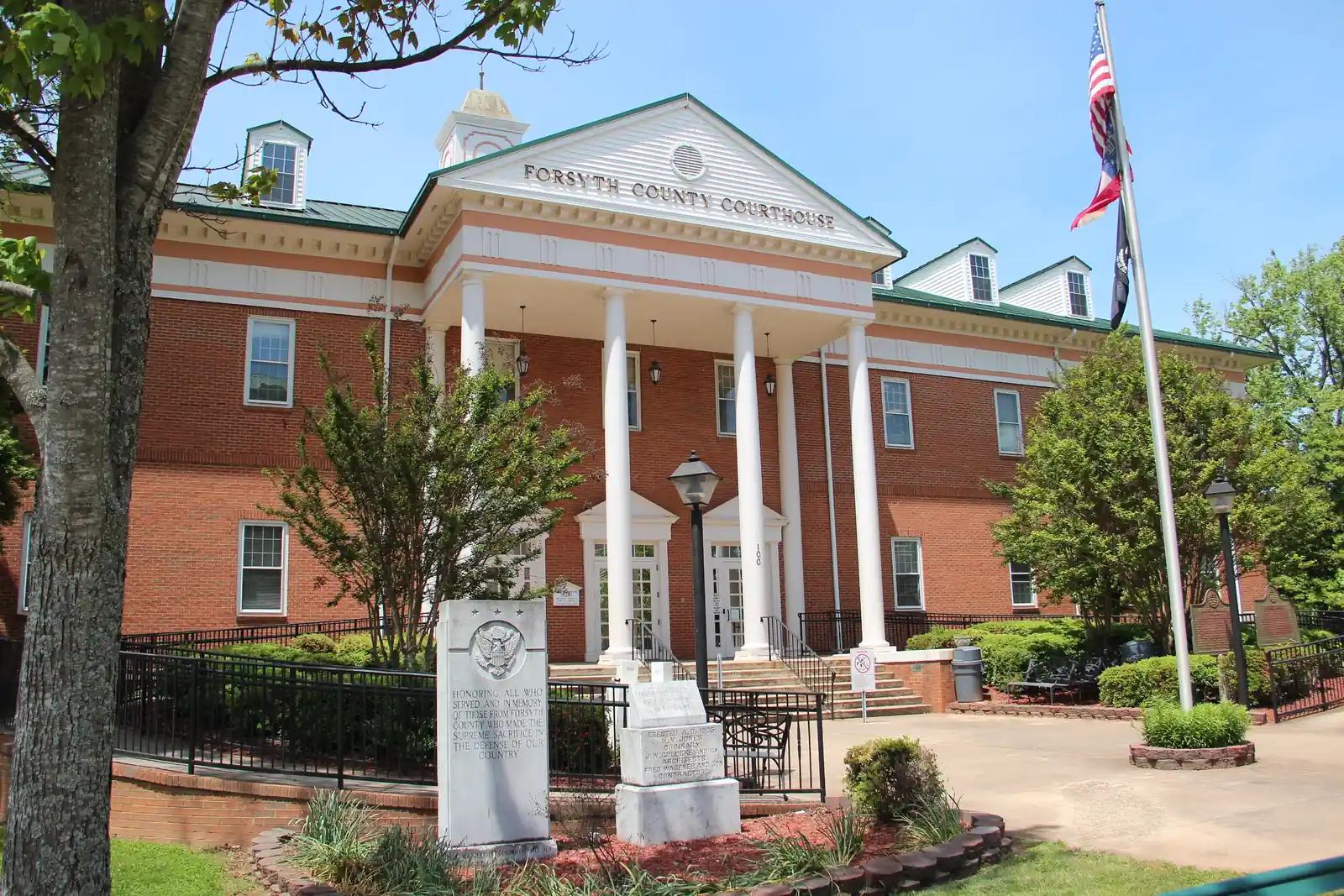 Two-storey red-brick Georgian-style building with a white columned portico and pediment lettered 'FORSYTH COUNTY COURTHOUSE', dormers and a cupola, a granite veterans' memorial ('Honoring all who served ...') in front, a tall flagpole with US and Georgia flags, a tree trunk framing the left edge. No people.