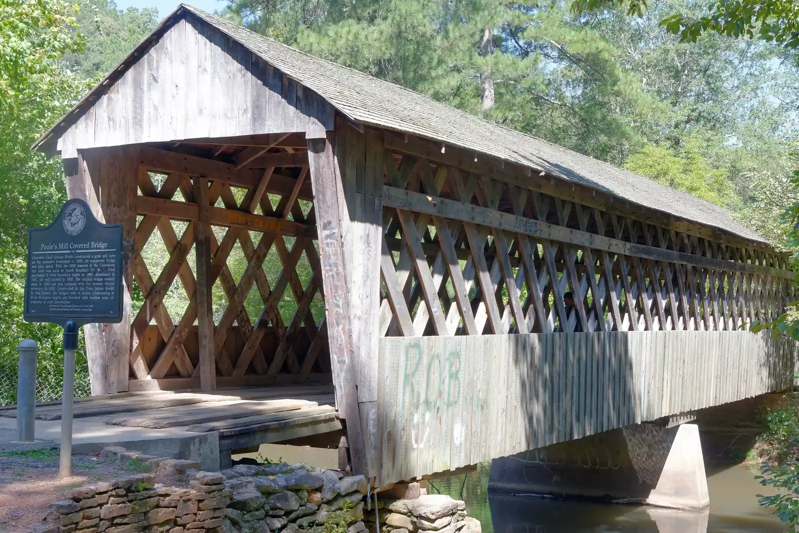 Sunlit three-quarter view of the weathered grey timber covered bridge with lattice (town-truss) sides and a shake roof, a Georgia Historical Society marker headed "Poole's Mill Covered Bridge" on a post at left, dry-stacked stone abutment in the foreground and the creek below; green spray-paint graffiti ('ROB') on the lower side boards; tall pines behind. No people.