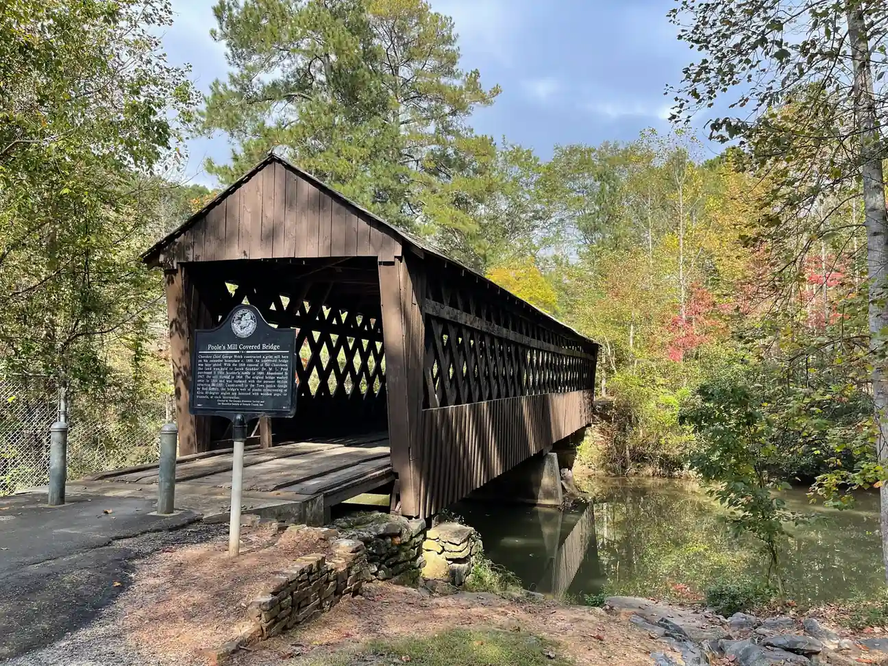 Autumn view of the dark-brown timber covered bridge (freshly stained, no graffiti) with lattice sides spanning Settingdown Creek, the historical marker on a post in front, steel bollards and an asphalt approach at left, stone wall, and trees turning red and gold along the creek with the bridge reflected in still water at right; blue sky with clouds. No people.