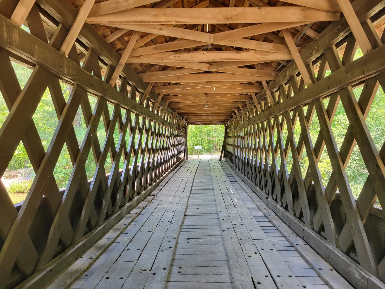 Symmetrical one-point-perspective view through the bridge interior: worn plank deck running to a bright opening, diagonal lattice trusses on both sides with green summer foliage glowing through, and the exposed roof framing overhead. A small interpretive sign is visible far away in the exit opening (not legible). No people.