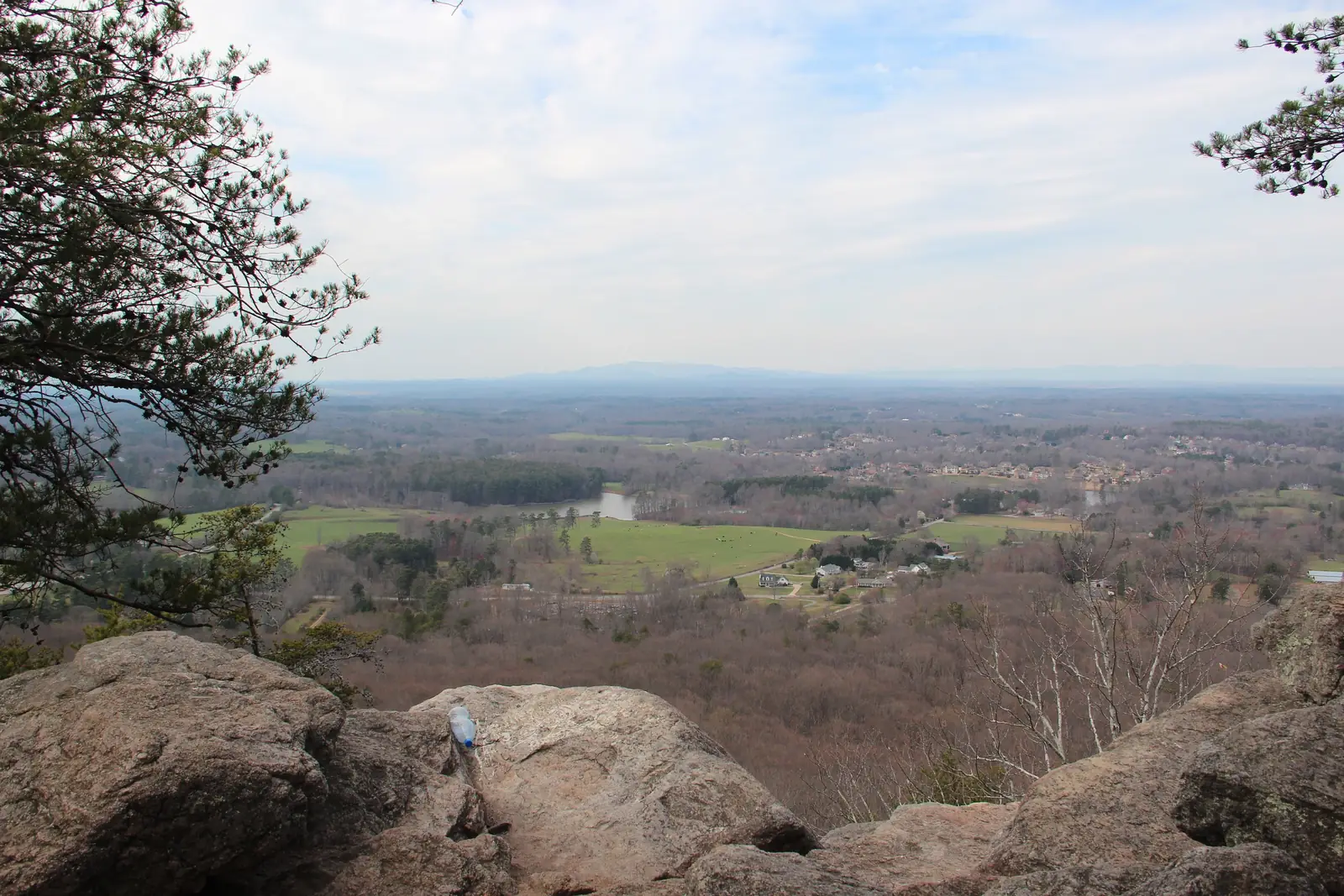 Early-spring view from the Indian Seats rock outcrop: grey granite boulders frame the bottom, a pine leans in at left, and below spreads a patchwork of bare winter woods, green pastures, a small farm pond and scattered homes and subdivisions toward the Blue Ridge silhouette on the horizon under a pale overcast sky. A small plastic water bottle sits on the rock at lower centre-left.
