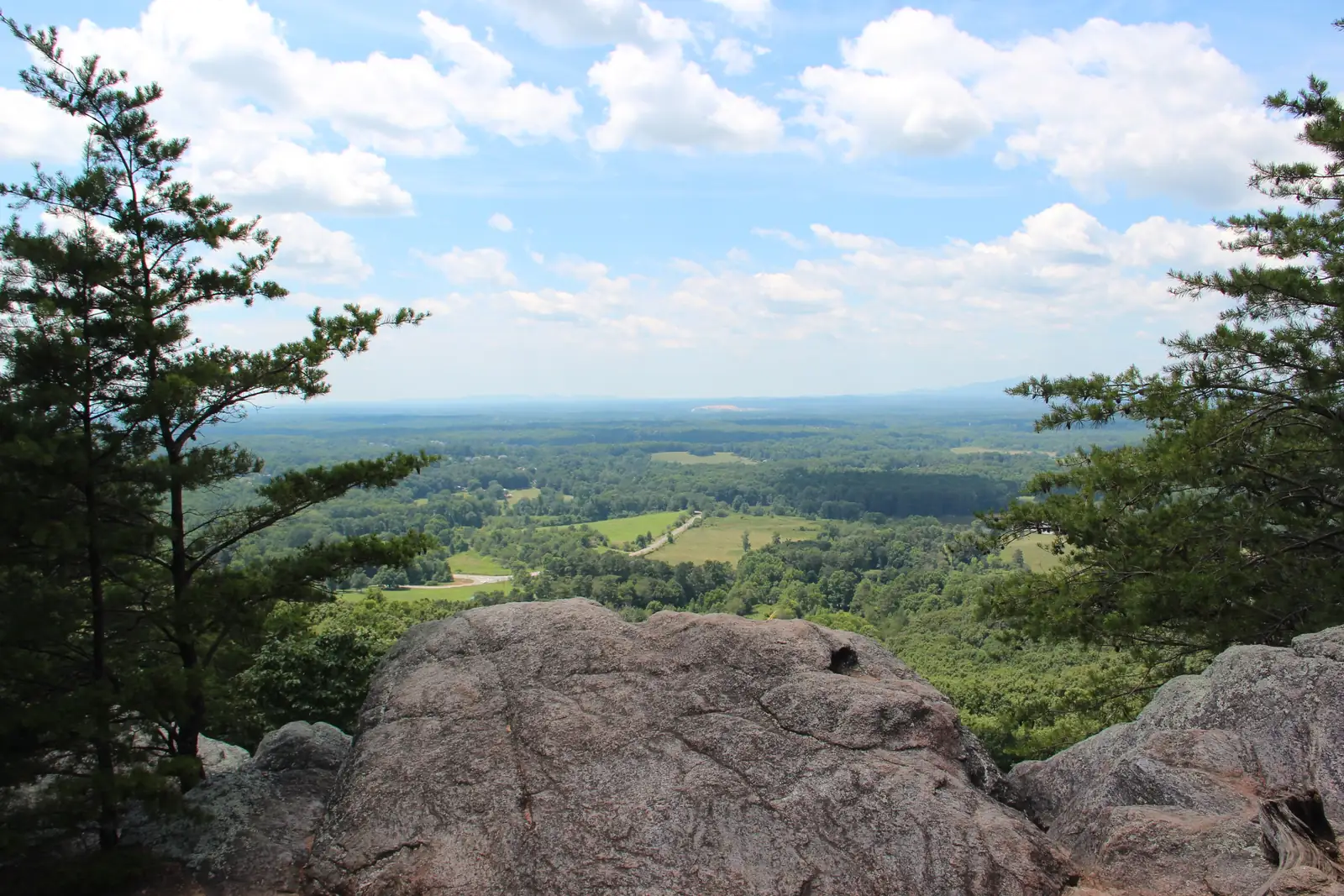 Summer view framed by two pines from a large pink-grey granite boulder on Sawnee Mountain: rolling green farmland with fields, tree lines and a winding road below, hazy blue ridges on the horizon, bright blue sky with puffy white clouds. No people, no signs.