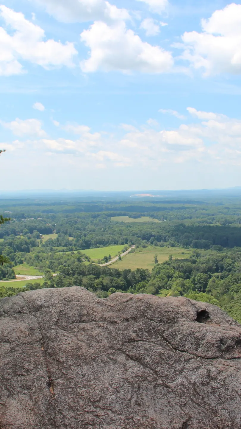 Summer view framed by two pines from a large pink-grey granite boulder on Sawnee Mountain: rolling green farmland with fields, tree lines and a winding road below, hazy blue ridges on the horizon, bright blue sky with puffy white clouds. No people, no signs.