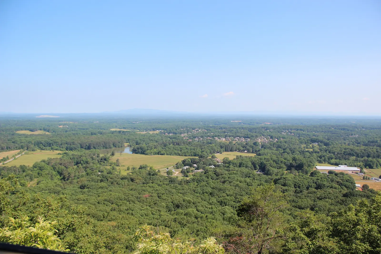 Bright summer panorama from Sawnee Mountain: deep green forest canopy in the foreground, pastures, a farm pond, long white agricultural barns at right and subdivisions in the middle distance, flat hazy horizon with a faint mountain outline, cloudless pale-blue sky filling the upper ~45%. A dark railing edge at the very bottom.