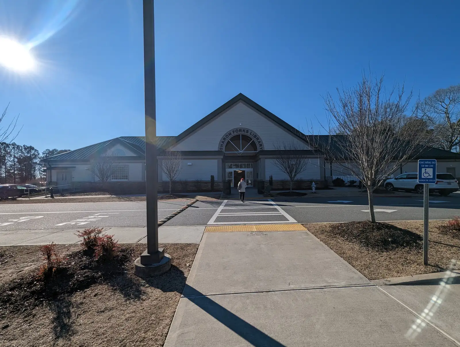 Winter view across a parking-lot crosswalk to the Sharon Forks Library: a pale gabled building with a fan window and 'SHARON FORKS LIBRARY' lettering over the entrance, bare young trees, a light pole in the foreground, a blue disabled-parking sign, parked SUVs at right and strong sun flare at top left. One person walking toward the door, seen from behind and too small to identify.