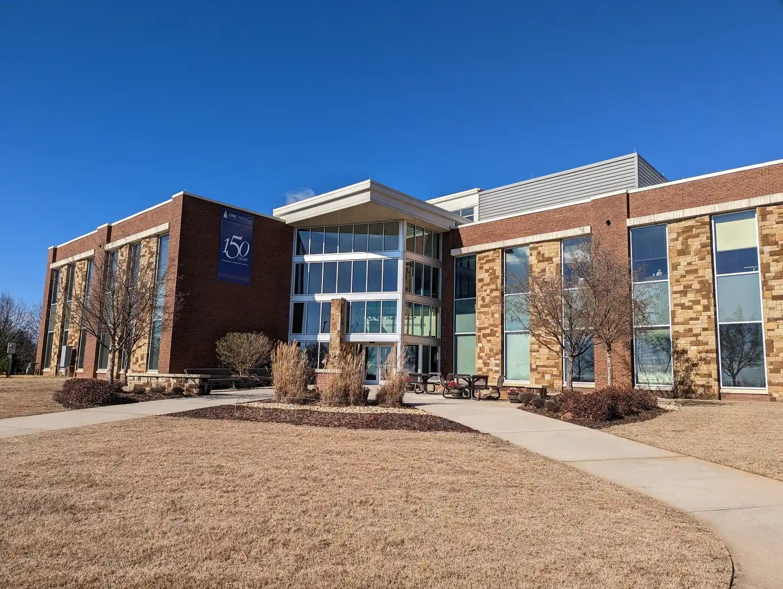 Two-storey brick, stone-veneer and glass academic building under a deep blue winter sky, a glazed entrance bay, a navy vertical banner reading '150 years' with the UNG logo, bare young trees, dormant tan lawn and a curving concrete walkway in the foreground. No people.