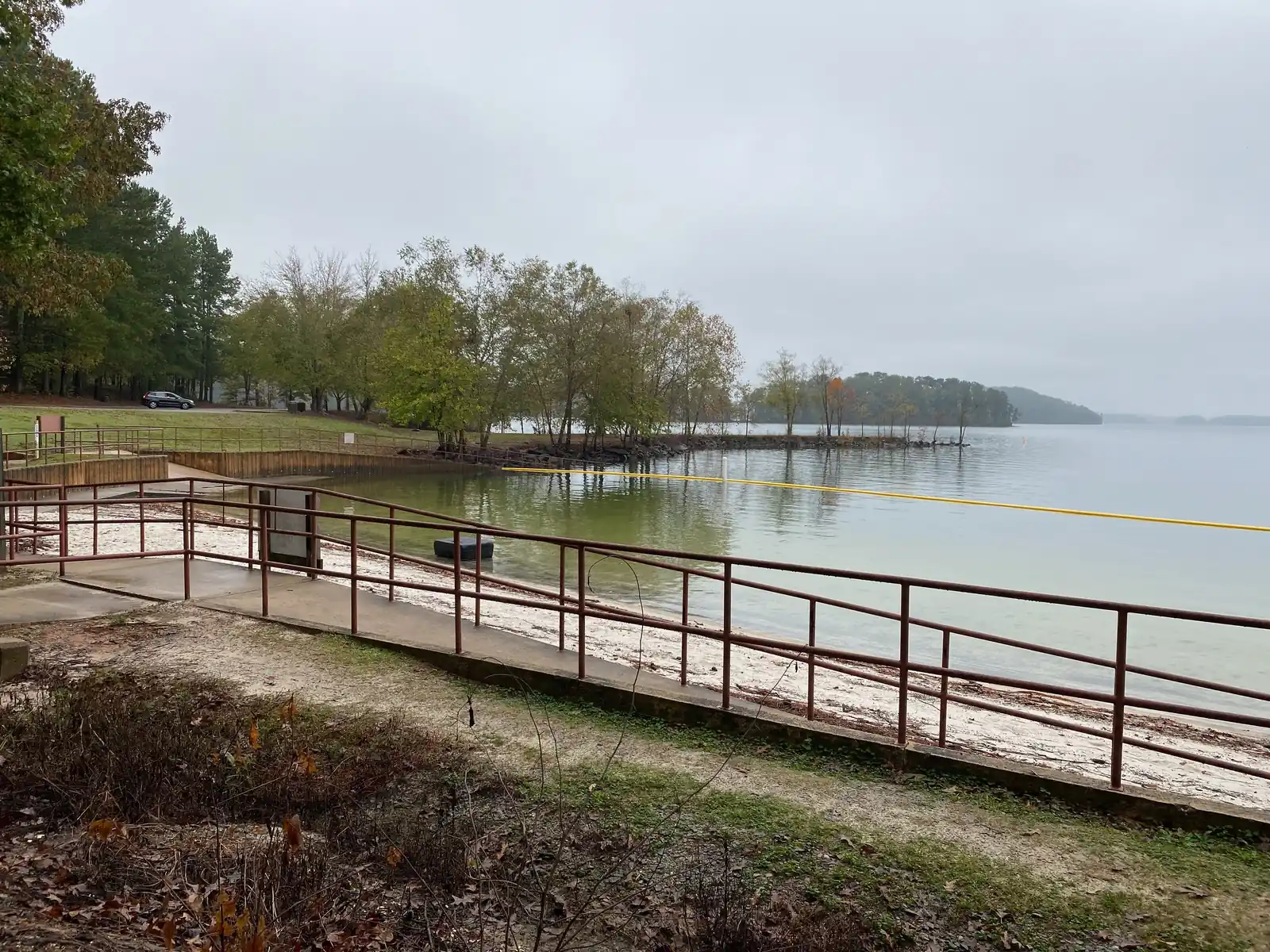 Overcast autumn morning at the West Bank Park swim beach: a brown metal railing and concrete ramp run down to a pale sand beach in the foreground, a yellow swim-area rope crosses calm milky-green water, a line of yellowing trees runs out along a low point, and a wooded headland fades into mist on the right. One dark parked car on the far lawn at left. No people, no legible signs.